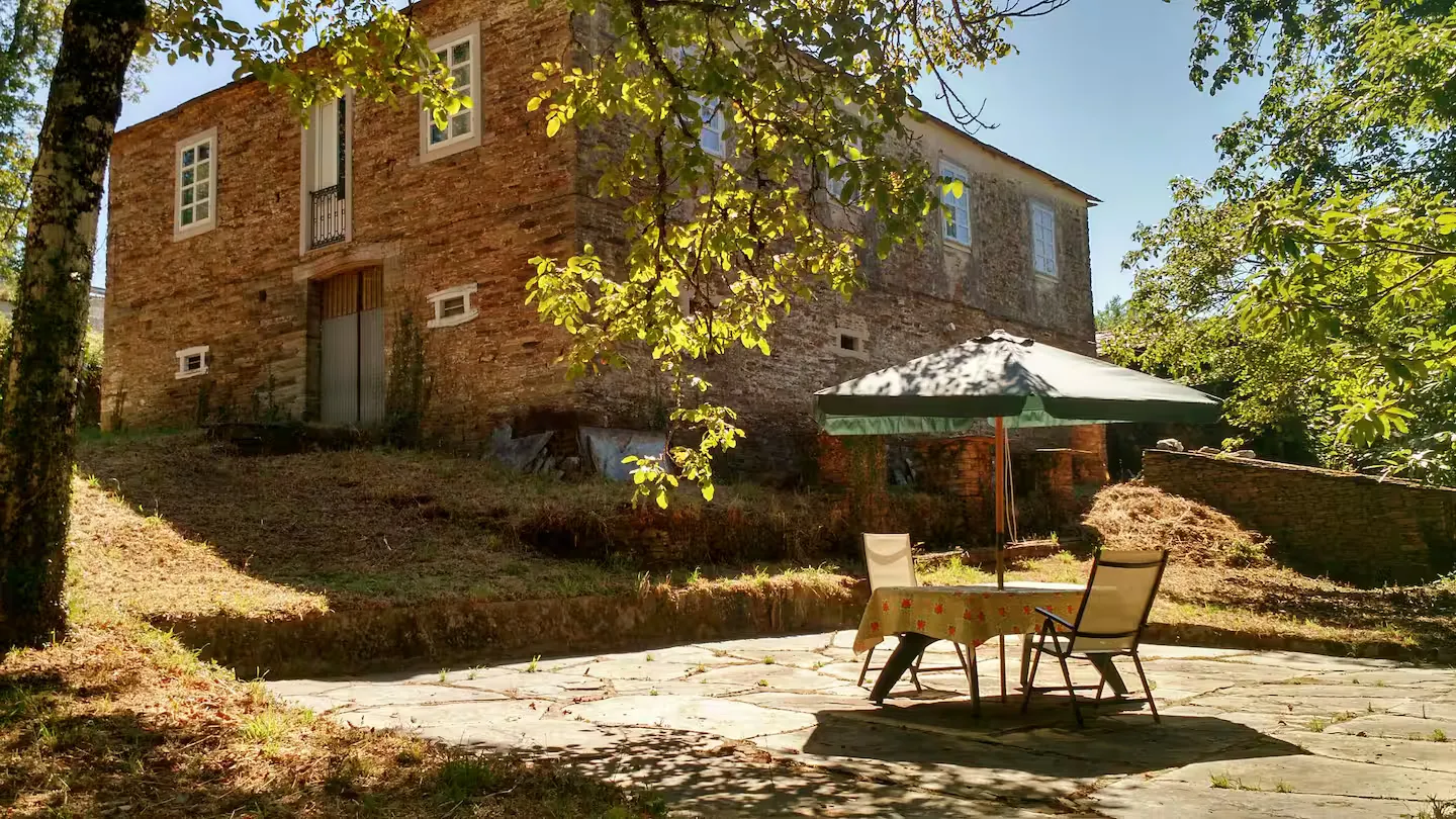 Fachada de piedra de Casa Moreda, con mesa y sombrilla en el patio bajo los árboles